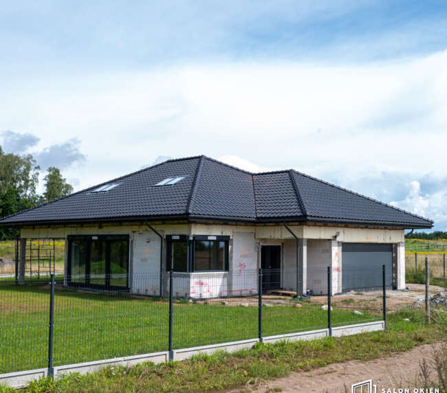 Dom parterowy z dużymi przeszkleniami, drzwiami i bramą garażową widziany zza ogrodzenia