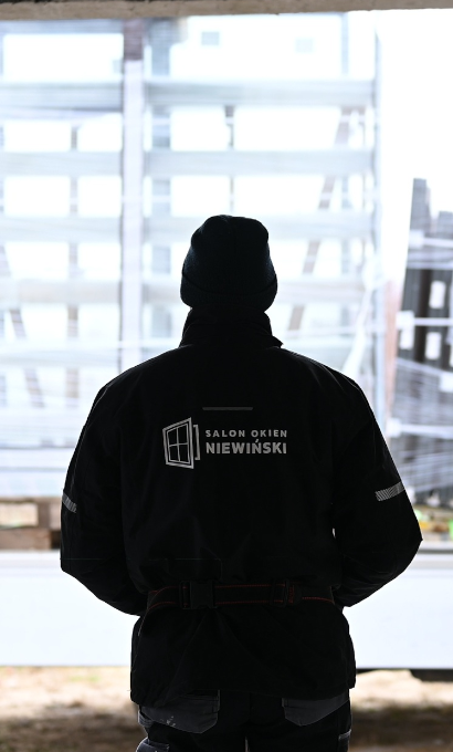 Monter w kurtce z logo Salonu Okien Niewiński stojący tyłem przed stojakami z oknami